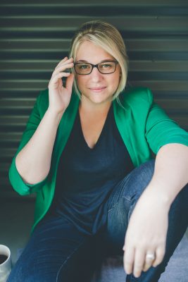 A blond woman with glasses crouching on the ground, wearing a green cardigan and jeans. 