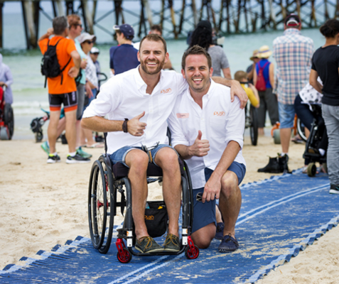 2 male appearing people on a blue beach mat, the left hand one is in a wheelchair, the right hand one is kneeling down. Both are wearing white shirts. 