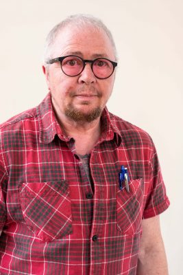 A male figure facing camera wearing glasses and a red check shirt. 