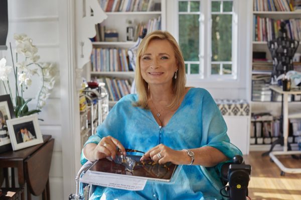 A blond woman in a power wheelchair smiling to camera, wearing a light blue top with floaty sleeves. 
