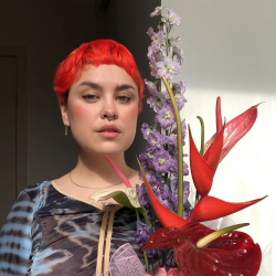 A person with bright red short hair, wearing a coloured top and holding a large bunch of purple and red flowers.