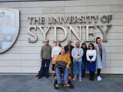 8 disabled people in front of a wall with University of Sydney in large letters. A person in the front is using a power wheelchair, others are standing behind him.