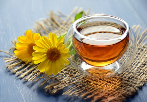 A brown cup of tea in a glass cup, sitting on a woven mat, next to it is a bright yellow flower. 