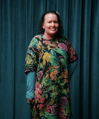 A smiling woman with dark curly hair facing camera, she is wearing a patterned frock and has a red face. 