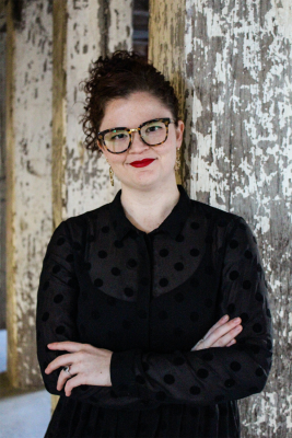 Lucy is facing camera with her arms crossed, she is wearing a black spotted outfit and has dark curly hair, glasses, red lipstick and gold earrings. 