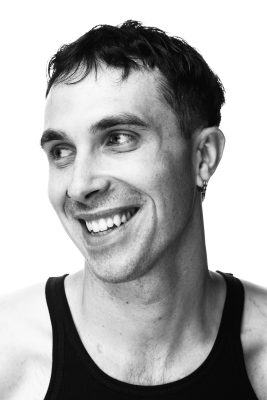 A black and white photo of a smiling man in a black singlet top. He is looking to left of the picture. 