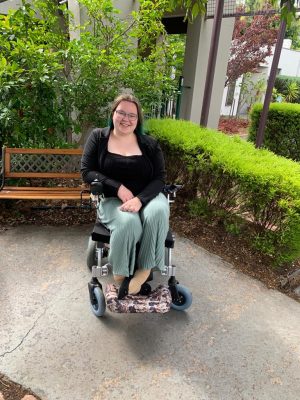 A woman with dark hair and glasses is smiling at the camera, she is sitting in a small power wheelchair in a garden. 
