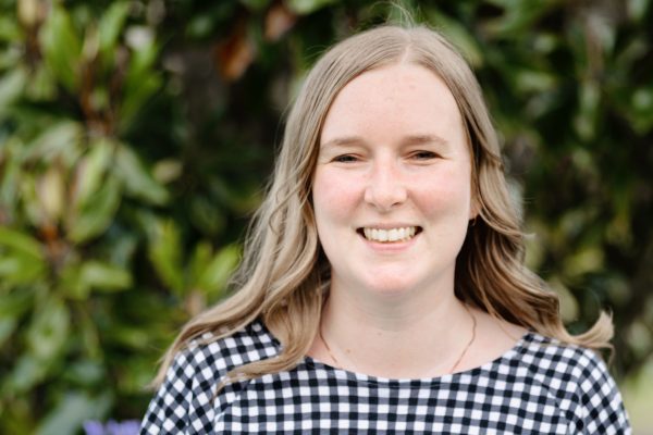 A woman with long blond hair smiling at the camera, she is wearing a check top and has trees behind her. 