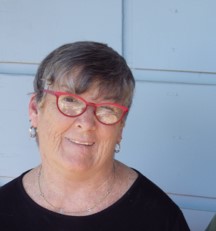 Karleen is facing camera, she has red glasses, grey hair and a black top. She is standing in front of a white wall. 