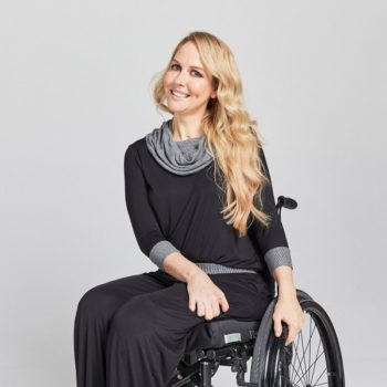 A woman sitting in a wheelchair, she is wearing long sleeved black and grey clothes and trousers and has long blond hair and is smiling. 