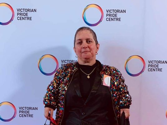 Margherita standing in front of a wall saying Victorian Pride Centre, she is wearing a sequin jacket and a big smile. 