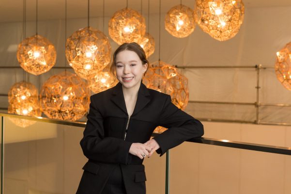 Natalia is leaning on a railing, she has dark hair and is wearing black clothes. There are big globe lights behind her. 