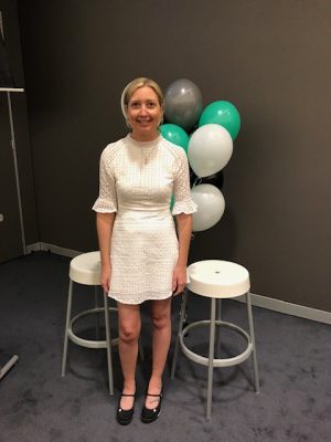 Sarah is smiling to camera, she has blond hair and is wearing a short white frock and is standing in front of balloons. 