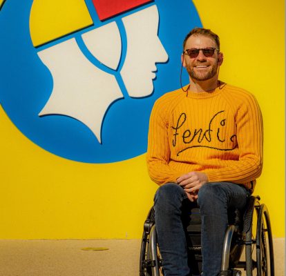 A smiling person in an orange top, they have dark hair and sunglasses and are sitting in a wheelchair in front of a lifesaving logo on a wall. 