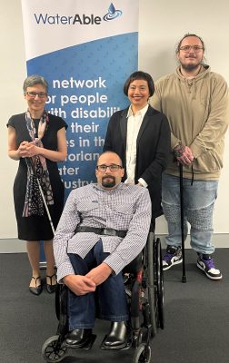A group of 4 people are in front of a blue and white WaterAble banner. One has a white cane, another is in a wheelchair, one is leaning on a cane. All are smiling. 