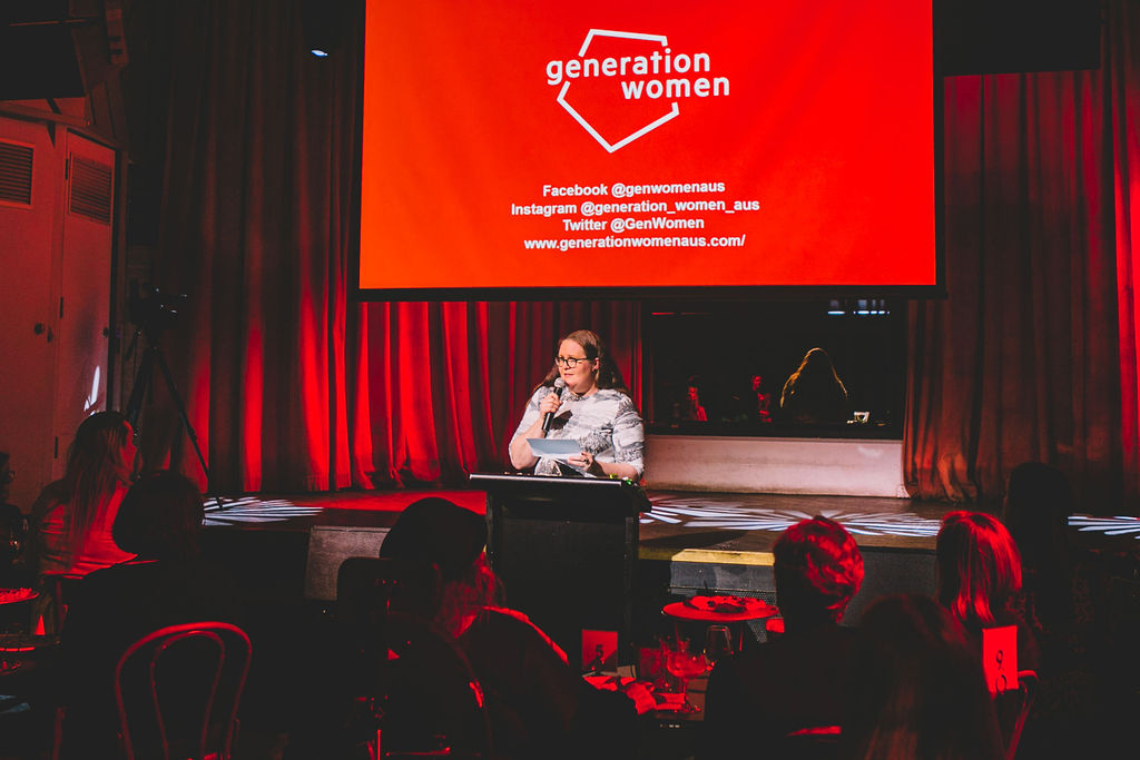 Morwenna standing and presenting at a lectern in front of a red screen that says 'Generation Women'.