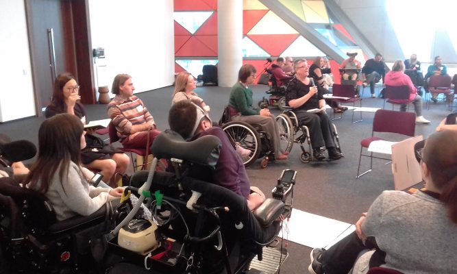 A group of disabled people in a circle at a conference. Some are in wheelchairs, they are talking together. 