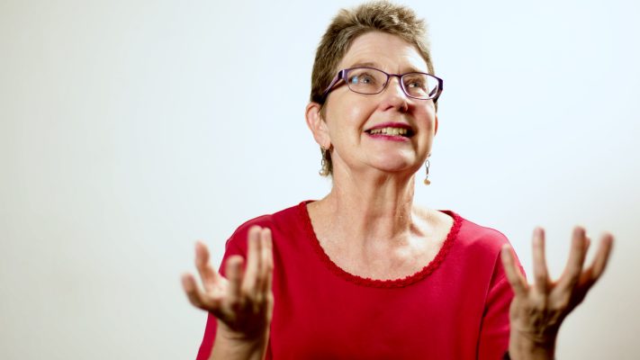 Annette is wearing a red tshirt and glasses, she is looking up to the right of picture and has her hands in the air at shoulder height.