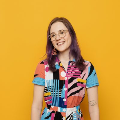 Ebe is against a yellow background, with shoulder length coloured hair, wearing tortoise shell glasses and a patterned top. She is smiling and looking to the left.