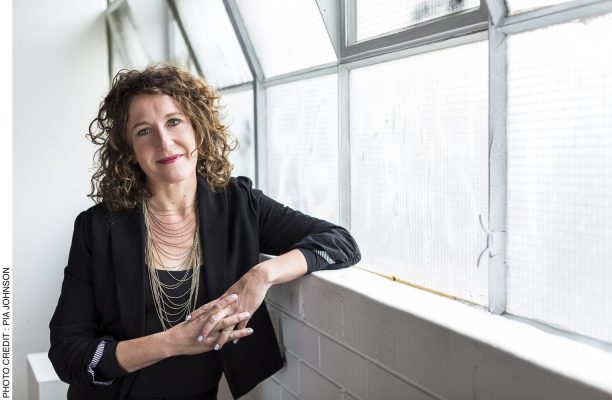 Jodee is leaning on a window sill wearing a black drop and fancy necklace. She has shoulder length curly brown hair.