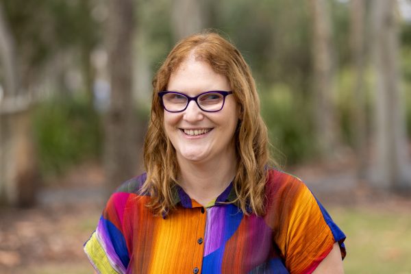 Katie has shoulder length red hair, a multi coloured top, and dark rimmed glasses. She has a big smile.