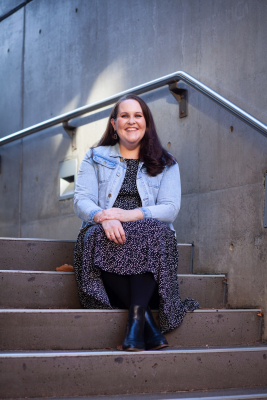 Morwenna is sitting on concrete steps, she is wearing a dress and denim jacket with a big smile and long dark hair.