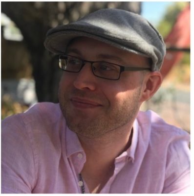 Aaron is wearing a pink shirt and a cap, he also has glasses on and is looking towards our left. He has short hair.