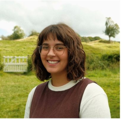 Chloe has shoulder length brown hair and glasses, she is smiling. Chloe is against a grass field background, she is wearing a brown top over a white long sleeved top.