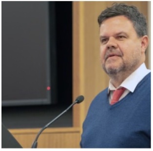 Damian is speaking at a lectern, he is wearing a blue jumper and red tie he has dark hair and a short beard.