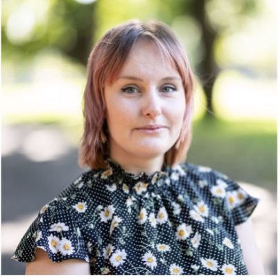 Danielle has blonde hair and is wearing a patterned black top. She is against a background of trees in a park.