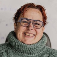 Fi is wearing a large greenish jumper with a big roll neck. They have short reddish hair and glasses.