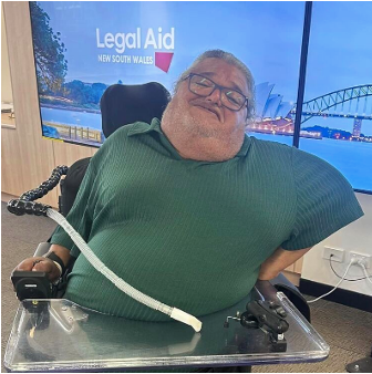 George is sitting in a power wheelchair and wearing a green tshirt. Behind him is a Legal Aid NSW picture with the Opera House and Harbour Bridge in it.