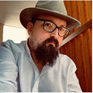 Jarrod is wearing a white shirt and a large hat, he has dark hair, glasses and a long beard.