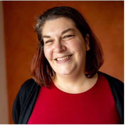 Karen has shoulder length dark hair and is wearing a red top and black cardigan. She is against a brown background.