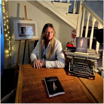 Kathie is sitting at a table with an old typewriter on it and a book. She is in front of an ornate staircase. She has long blonde hair and is wearing a white jacket.