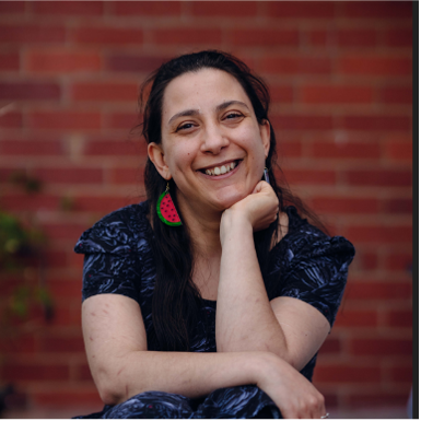 Laura is sitting down low and is resting her arms on her knees. She has dark hair, big red earrings and is wearing a blue dress.