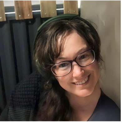 Linda is wearing glasses and a black top. They have long dark hair and headphones on.