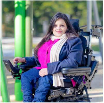 Nabila is sitting in a large power wheelchair, she has on a pink top underneath a blue coat. She has long dark hair and bright lipstick.