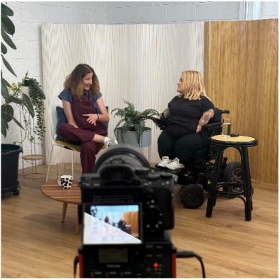 Two disabled women are sitting talking to each other with a camera filming them. One is sitting is a power wheelchair the other is talking and gesticulating. There is a pot plant to the left.