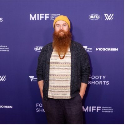 Ramas is wearing a black relaxed shape suit and a yellow beanie. He has a large red beard. He is standing in front of a Melbourne International Film Festival media wall with logos of the AFL and Footy Shorts on it.