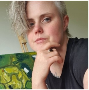 Sarah has short blond hair and is looking at us with her chin resting on her right hand. She is wearing a black top and is in front of a green coloured artwork on an easel.