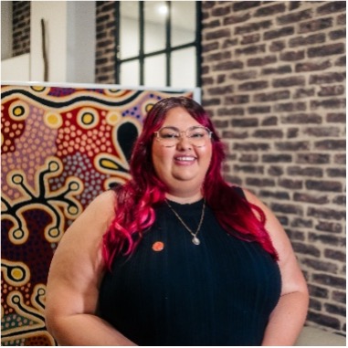 Tahlia has red hair, a black top and glasses and is standing in front of a First Nations artwork and a brick wall. She is smiling!
