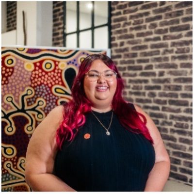 Tahlia has long red coloured hair and glasses, she is wearing a black top. She is in front of an Indigenous artwork and a brick wall.