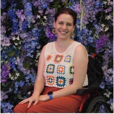 Tessa is wearing a crochet white top and brown trousers, she is sitting in a red manual wheelchair. She has short dark hair and large yellow earrings.