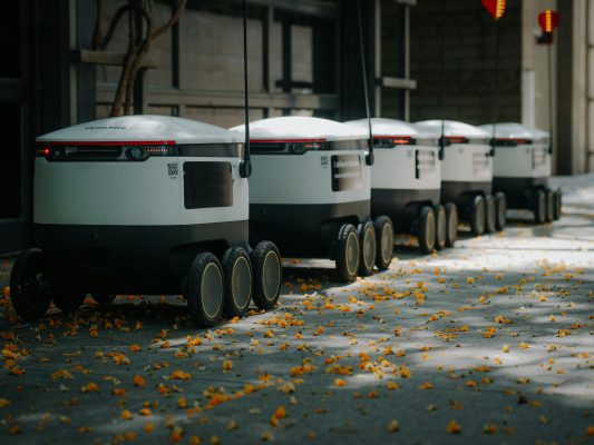 A line of boxy worker type robots with 6 wheels following each other.