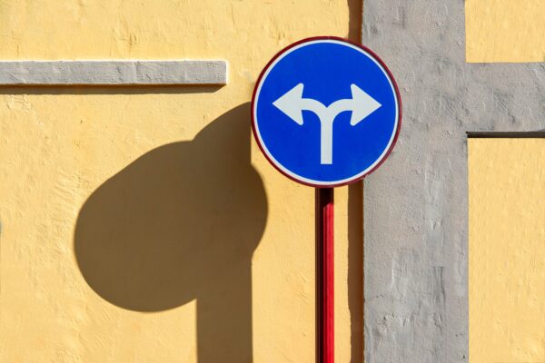 A blue road sign against a yellow wall, the sign has a white arrow pointing to both left and right. 